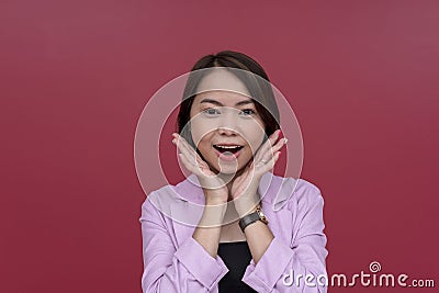 A young asian woman looking thrilled with hands on her cheeks. Wearing a pink blazer and black tube top. Studio shot with burgundy Stock Photo