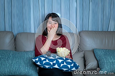 Young asian woman watching television suspense movie or news looking happy and funny and eating popcorn late night at home living Stock Photo