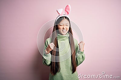 Young asian woman wearing cute easter bunny ears over pink background excited for success with arms raised and eyes closed Stock Photo