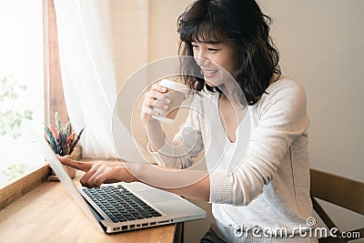 Young Asian woman working with laptop computer and drinking a cup of coffee. A woman with smiley face working from home. Social Stock Photo