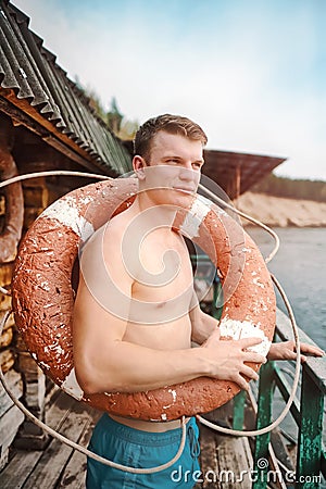 This is a young athletic guy in shorts and bare-chested standing with an old lifebuoy. Stock Photo