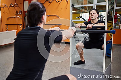 Young athletic man in black sportwear with bar of barbell flexing muscles in gym in front of the mirror. Stock Photo