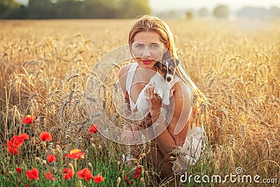 Young athletic woman, kneeling, holding Jack Russell terrier puppy on her hands, some red poppy in foreground and sunset lit wheat Stock Photo