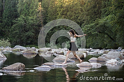 Young attractive brunette woman hopping over rocks beside a pristine river in an evergreen forest. Stock Photo
