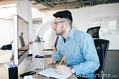 Young attractive dark-haired man in glassess is working with a computer at his workplace in office. He wears blue shirt Stock Photo