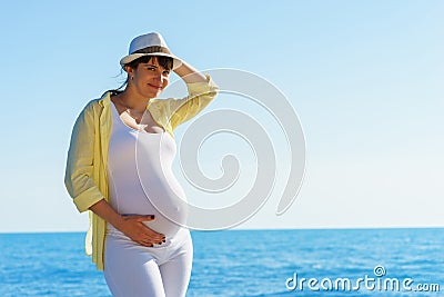 Young attractive european pregnant woman in yelow shirt and white hat is staying at the shore of tranquil blue sea and holding tum Stock Photo