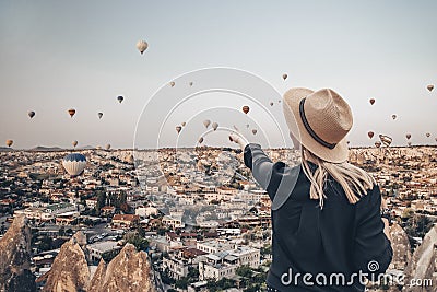 Young attractive girl in a hat stands on the mountain with flying air balloons on the background. Finger pointing girl Stock Photo