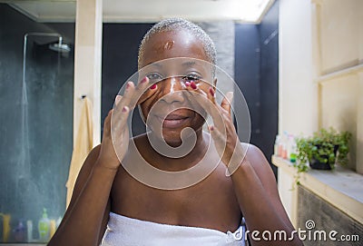 Young attractive and happy black african American woman at home bathroom looking on toilet mirror applying face beauty moisturizer Stock Photo