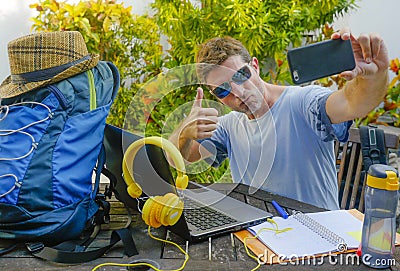 Young attractive and happy digital nomad man working outdoors with laptop computer cheerful taking selfie pic with mobile phone Stock Photo