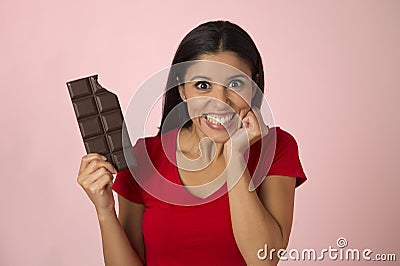 Young attractive and happy hispanic woman in red top smiling excited eating chocolate bar on pink background Stock Photo