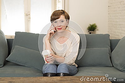 Young attractive and happy red hair woman sitting at home sofa couch drinking coffee talking on mobile phone relaxed in communicat Stock Photo