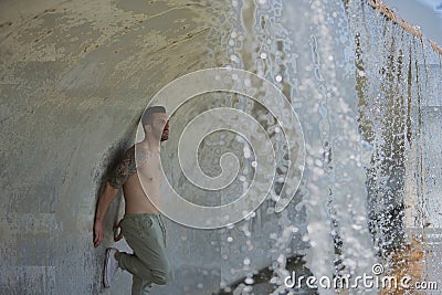Young, attractive and Hispanic man, shirtless and with a muscular body, leaning against a wall under a bridge where a waterfall Stock Photo
