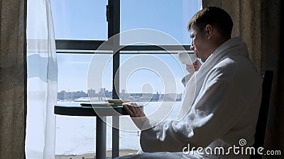 A young, attractive man sitting at a table by the window drinking tea, coffee and writing an SMS message on a mobile Stock Photo
