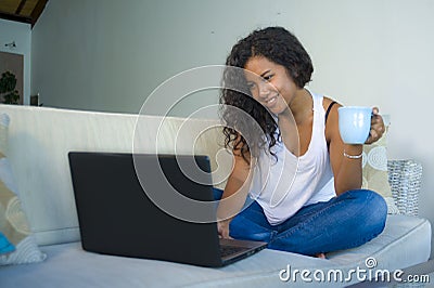 Young attractive and relaxed hispanic student woman sitting at home sofa couch networking with laptop computer drinking coffee or Stock Photo