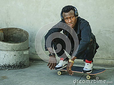 Young attractive and serious black afro American man squatting on skate board at grunge street corner looking cool posing in Stock Photo