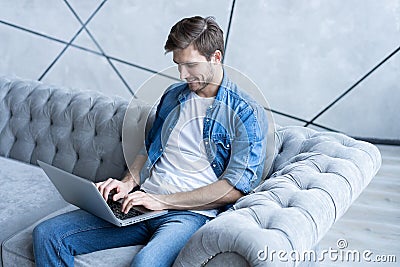 Young attractive smiling guy is browsing at his laptop, sitting at home on the gray sofa at home, wearing casual outfit. Stock Photo