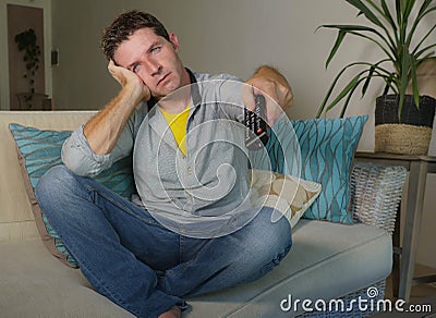 Young attractive unhappy man in casual clothes at home bored and frustrated watching tv movie on living room sofa couch holding re Stock Photo