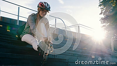 Young attractive woman adjusting rollerblades while sitting on the stairs in the skatepark. Direct sunshine Stock Photo