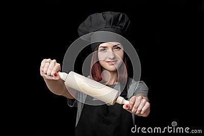 Young attractive woman chef in black uniform holds rolling pin on black background. Focus on rolling pin Stock Photo