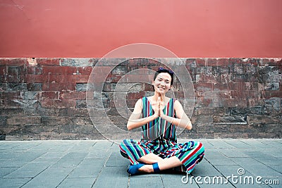 Young attractive woman sitting in yoga pose and smiling outdoor over red wall, image toned. Chinese girl amuses, summertime. Stock Photo