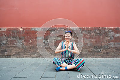 Young attractive woman sitting in yoga pose and smiling outdoor over red wall, image toned. Chinese girl amuses, summertime. Stock Photo