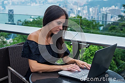 Young attractive woman typing or working on laptop sitting in rooftop cafe with city view. Stock Photo