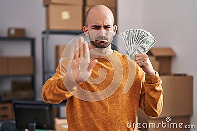 Young bald man with beard working at small business ecommerce holding money with open hand doing stop sign with serious and Stock Photo