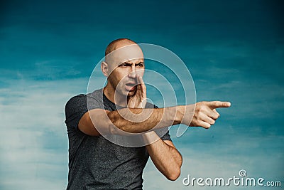 A young bald man in a gray T-shirt on a blue background points a finger. Stock Photo