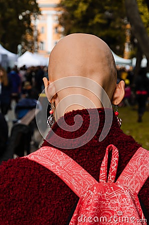 Young bald woman with long earrings. Dressed in a burgundy coat with a pink backpack. Back view. Outdoors Editorial Stock Photo