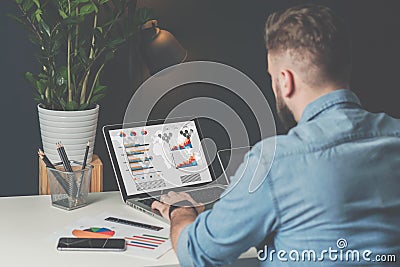 Young bearded businessman in denim shirt is sitting in office at table and is using laptop with charts, graphs Stock Photo