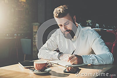 Young bearded businessman sits in cafe at table and writes in notebook.On table tablet computer, smartphone.Man is Stock Photo