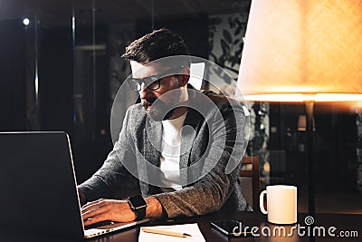 Young bearded businessman sitting at wooden table with lamp in loft office at night. Project manager working process and typing te Stock Photo