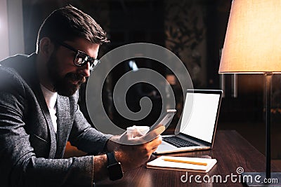 Young bearded businessman using phone while sitting by the wooden table in modern office at night. People working mobile devices Stock Photo