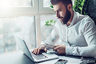 Young bearded businessman in white shirt is sitting at table and working on laptop. Freelancer works at home. Student Stock Photo