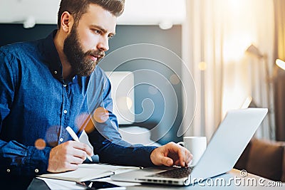 Young bearded businessman works on computer, takes notes in notebook, analyzes information, makes business plan. Stock Photo