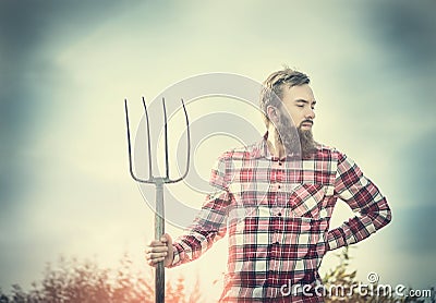 Young bearded farmer in red checkered shirt with old pitchfork sky nature backgrund, toned Stock Photo