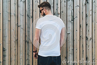 Young bearded hipster man dressed in white t-shirt and sunglasses is stands outdoor against wood wall. Mock up. Stock Photo