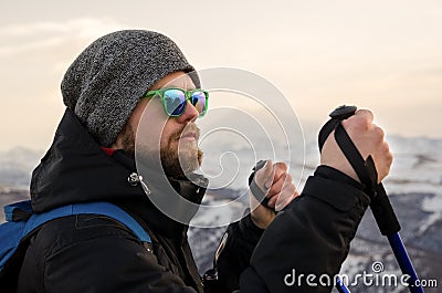 Young bearded hipster wearing a hat and sunglasses with Nordic walking sticks and a backpack moves in the mountains at Stock Photo