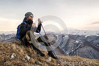 Young bearded hipster wearing a hat and sunglasses with Nordic walking sticks and a backpack moves in the mountains at Stock Photo