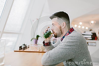 Young bearded man in cafe. He dressed in gray pullover and plaid shirt. Sits at long table near window. Man drinks Stock Photo