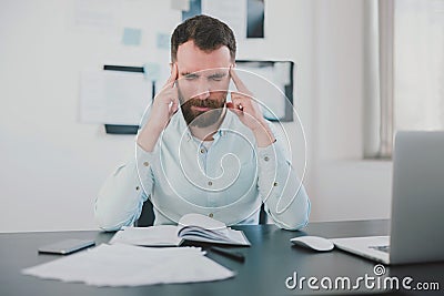 Young bearded man looking miserable suffering extreme headache at work while studying business project in his modern office, holds Stock Photo