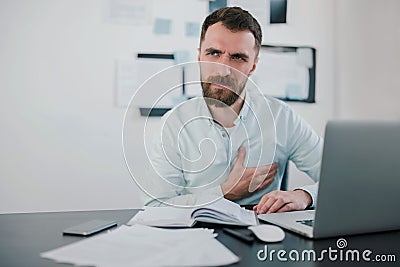 Young bearded man looking miserable suffering extreme heartache at work while studying business project in his modern office, Stock Photo