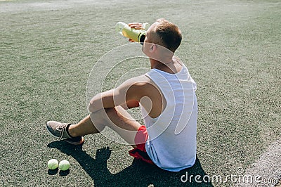 Young bearded man sitting on basketball court outdoors with ball holding bottle drinking electrolytes drink. Stock Photo