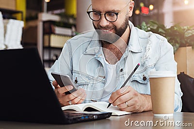 Young bearded man trendy glasses sits cafe in front of laptop computer, uses smartphone, takes notes in notebook Stock Photo