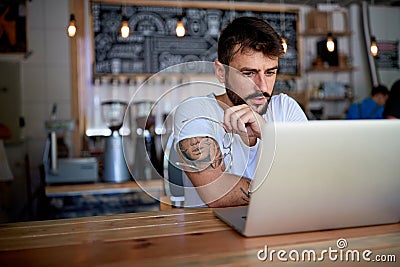 Young bearded tattooed guy in white t-shirt sitting behind the table in bar Stock Photo