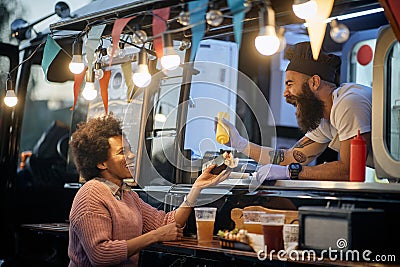 Young beardy caucasian employee in fast food laughing, adding a mustard in a sandwich to a female afro-american customer Stock Photo