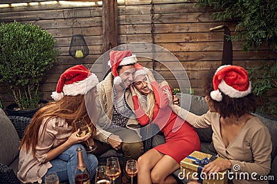 Young beardy guy hugging attractive blonde at Christmas party, wearing santa hat with other friends gathered Stock Photo