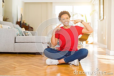 Young beautiful african american woman sitting on the floor at home smiling confident showing and pointing with fingers teeth and Stock Photo