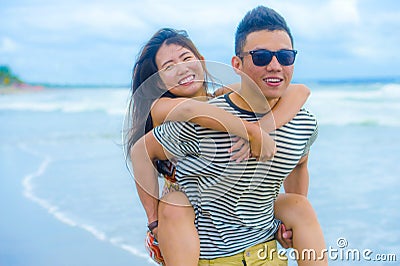 Young beautiful Asian Chinese couple with boyfriend carrying woman on her back and shoulders at the beach smiling Stock Photo