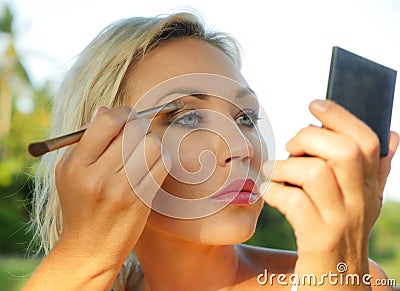 Young beautiful and attractive blond woman with blue eyes retouching makeup with brush applying eyeshadow holding small mirror in Stock Photo